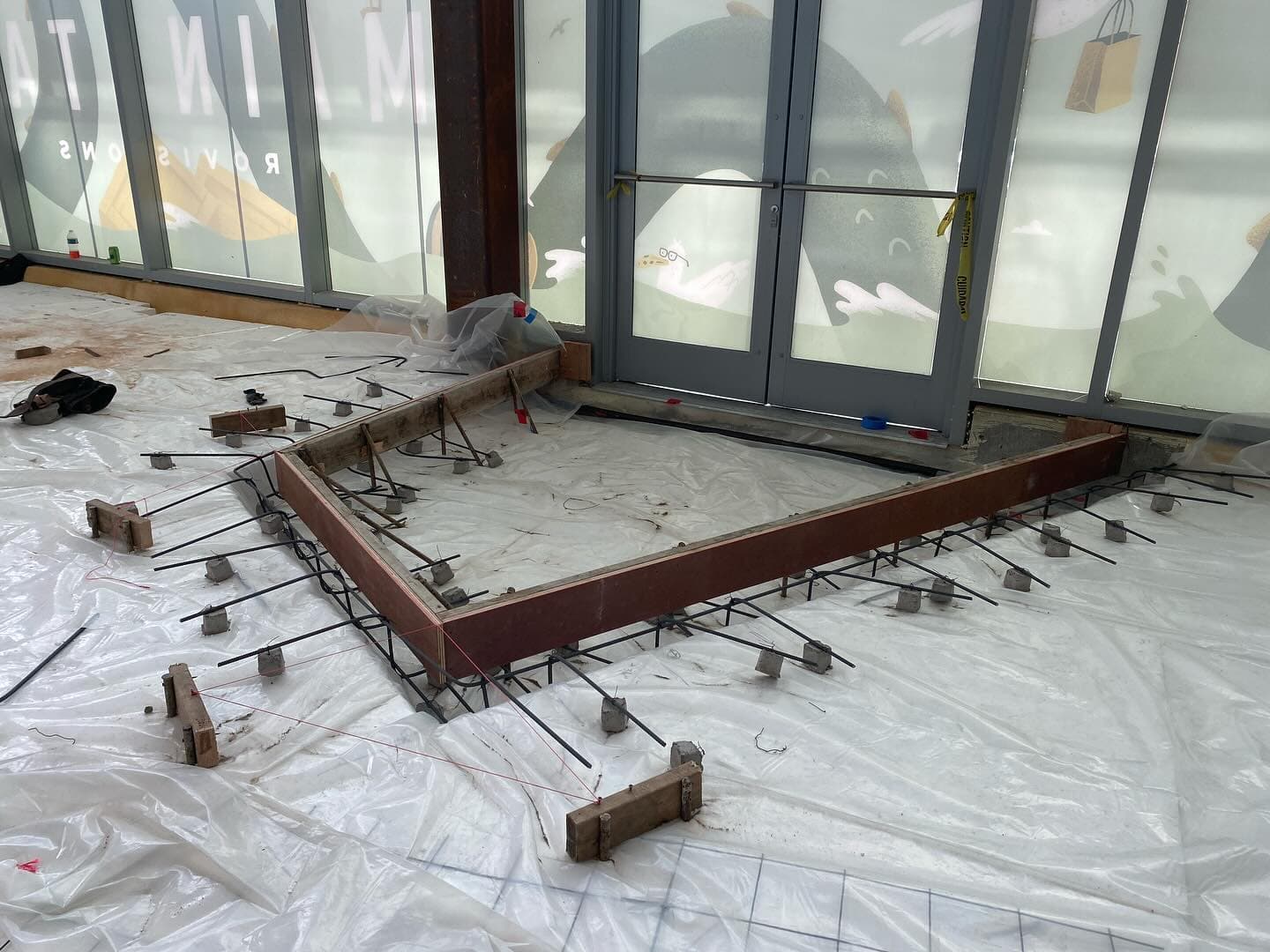 A raised slab formed in wood with rebar on chairs, set inside a storefront with plastic sheeting protecting the floor around it.