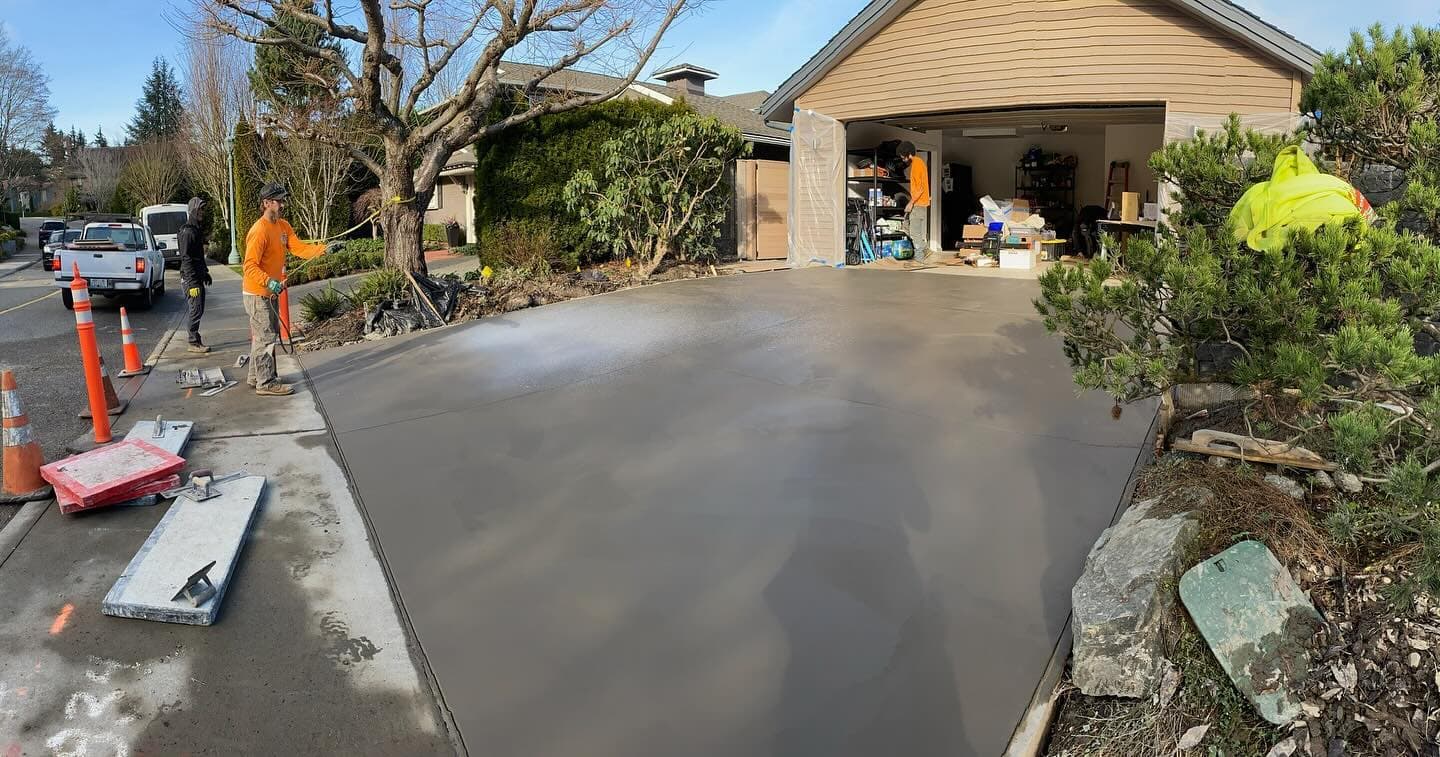 Three crew members placing and spreading concrete over rebar for a residential driveway in Seattle, the house behind them wrapped in plastic.