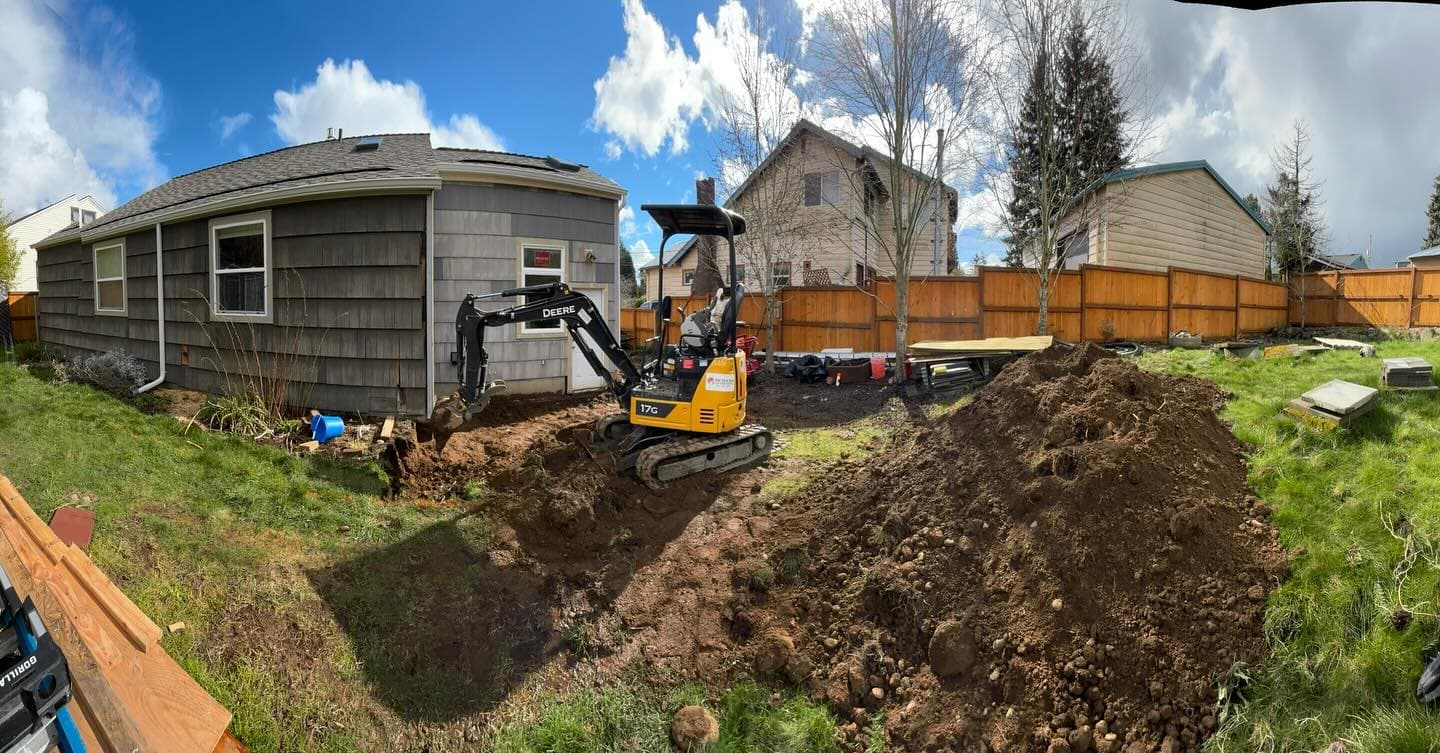 A mini excavator digging out a backyard beside a grey house, a pile of soil to the right and a new fence behind.