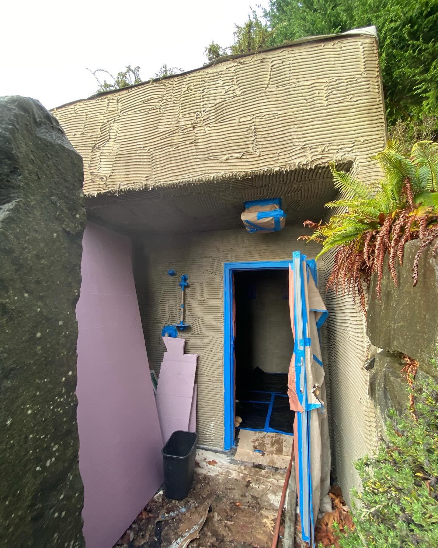 The entrance to an underground sauna and shower built into a hillside, its shotcrete walls hand-carved with horizontal ridges, the doorway taped off for staining.