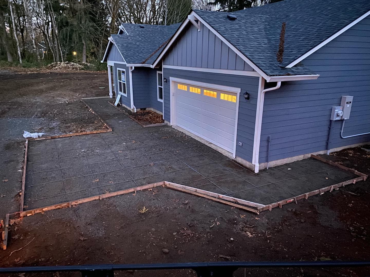 Driveway and entryway forms with rebar laid out in front of a new blue house at dusk, garage lights on.