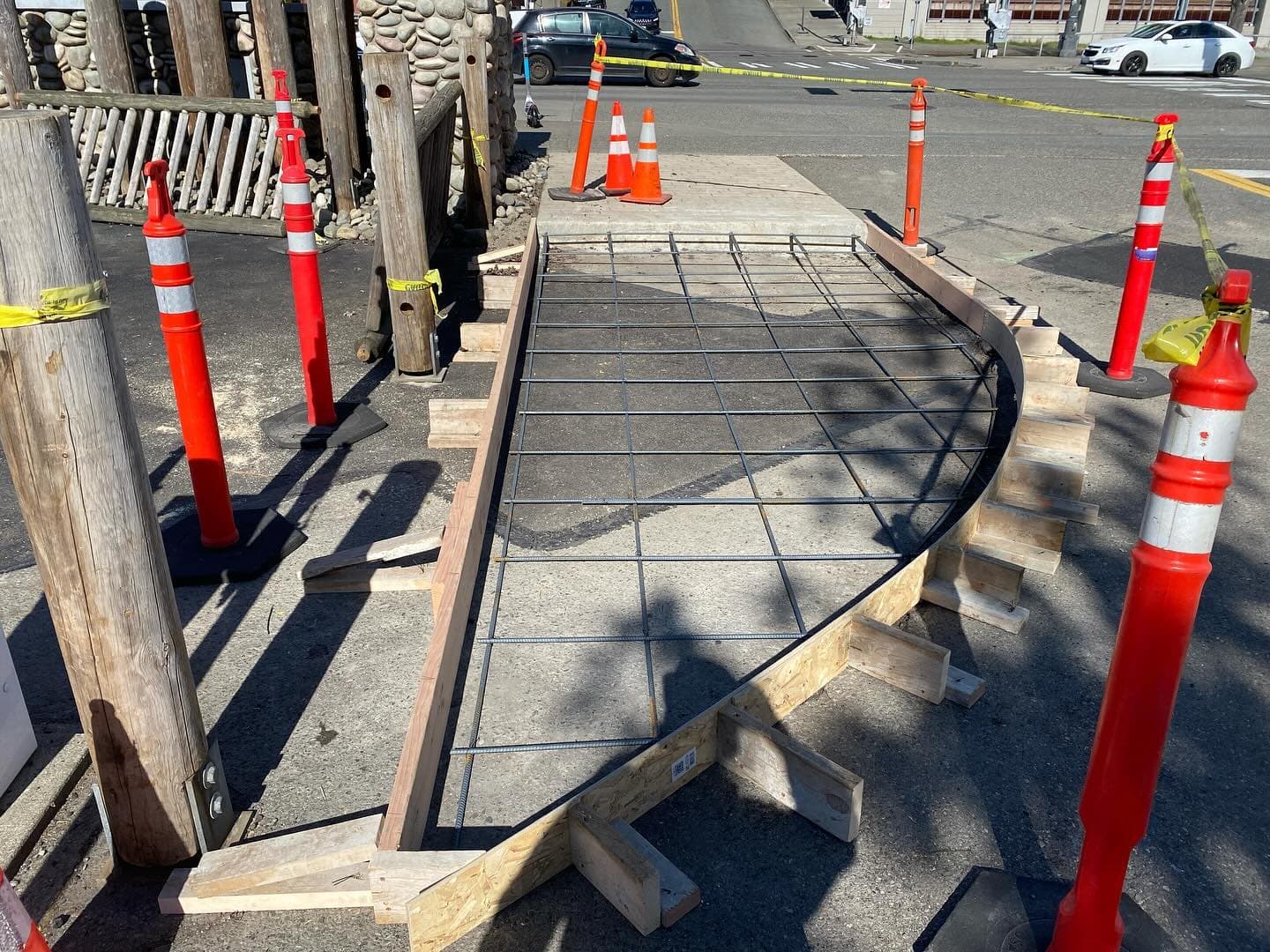 A curved sidewalk section formed with lumber and laid with a rebar grid, ringed by orange traffic delineators on a city street.